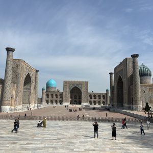 Panoramic view of Registan Square in Bukhara, Uzbekistan. The image features three intricately decorated madrasas (Islamic schools) flanking a large plaza. The central facade of Ulugbek Madrasa dominates the scene, with its tall portal and turquoise mosaic tiles. Kalyan Minaret, a towering brick minaret, stands to the right.